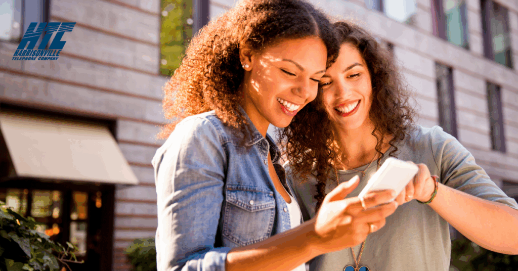 Two young women smile while looking at a smartphone together outside of a building - possibly watching their favorite shows.