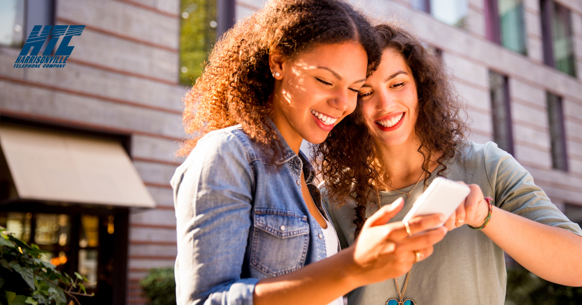Two young women smile while looking at a smartphone together outside of a building - possibly watching their favorite shows.